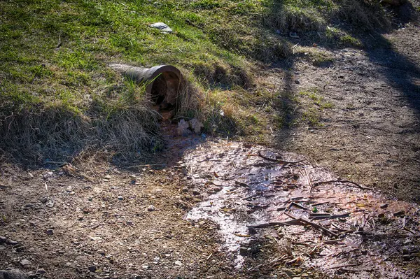 Paslı drenaj borusu kirlenmiş suyu toprağa sızdırarak kirlilik yaratır. Turuncu renkli akıntılar, endüstriyel atıkların neden olduğu çevresel hasarı vurguluyor.