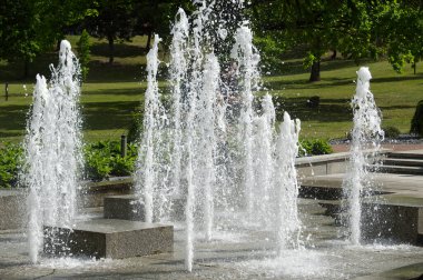 Dinamik bir fıskiyenin büyüleyici su fıskiyeleri serin bir parkta yeşilliklerle çevrili. Güneşli bir günde modern özelliklerin yanında doğanın güzelliğini gösteren sakin bir şehir manzarası..