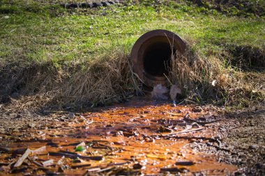 Aşınmış drenaj borusu kirlenmiş suyu toprağa sızdırır ve kirlilik yaratır. Bulanık turuncu akıntılar doğal ortamda endüstriyel atıkların neden olduğu çevresel hasarı vurguluyor.