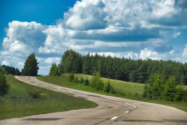 Kıvrımlı bir yol, gür, yeşil bir arazide, tüylü beyaz bulutlarla dolu parlak mavi bir gökyüzünün altında kıvrılır. Ağaçlar yol ve ufuk çizgisinde.