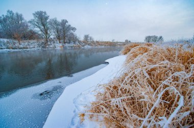 Huzurlu bir kış harikalar diyarı, güneş ufukta beliriyor, donmuş nehre sıcak altın bir ışık tutuyor, huzur ve doğal güzelliğin resimli bir sahnesini oluşturuyor. Kış Görkemi