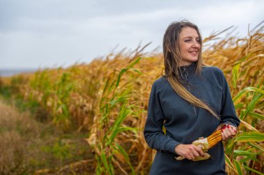 Sonbahar temalı fotoğraf çekimi bir kız ve bir mısır kulağıyla. Kız kalın gri bir elbise giyiyor. Arkadaki renkli ağaçlar