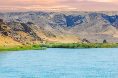 Huzurlu bir nehrin üzerinde günbatımının sakinliği. Görkemli dağların ve dingin bir ortamda yemyeşil ağaçların güzelliğinin tadını çıkar. Kendinizi bu manzaranın doğal güzelliğine daldırın..