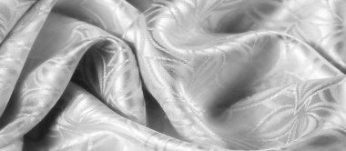 Akan gümüş-beyaz kumaşın zarif yakın plan çekimleri. Karmaşık, parlak desenleri var. Kumaş pürüzsüz, ipeksi bir doku sergiliyor, ince bir parıltı içindeki ince yaprak benzeri motiflerle vurgulanıyor..