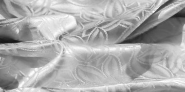 Pürüzsüz, lüks beyaz saten kumaşın yakın plan görüntüsü. Karmaşık çiçek desenleri var. Dokusunu ve detaylarını göstermek için yavaşça buruşmuş. Parlak yüzey ışığı yansıtır, narin bir parlaklık yaratır.