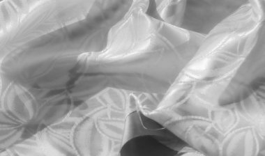 Pürüzsüz, lüks beyaz saten kumaşın yakın plan görüntüsü. Karmaşık çiçek desenleri var. Dokusunu ve detaylarını göstermek için yavaşça buruşmuş. Parlak yüzey ışığı yansıtır, narin bir parlaklık yaratır.