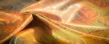 Işıldayan, çiçek desenli, altın, turuncu ve yeşil renkli kumaşların yakın çekimi. Karmaşık tasarım zarif kıvrımları ve dokuları vurguluyor. Yumuşak, aydınlık bir bitiş ile..