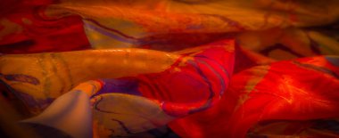 Sanatsal girdapları ve mor aksanları olan parlak turuncu ve kırmızı bir kumaş. Işığı yakalıyor ve soyut, akışkan bir tasarım yaratıyor..