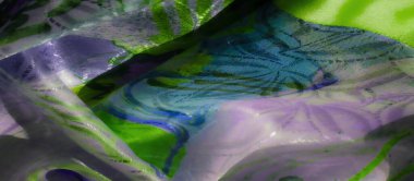 İç içe geçmiş yeşil, lavanta ve mavi tonlarda canlı ve soyut bir desen. Kumaş dokusu yumuşak ışık yansımaları ve keskin çizgilerle katmanlı bir etki yaratıyor..