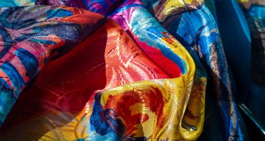 Mavi, sarı ve kırmızıları harmanlayan soyut suluboya desenleri olan canlı, kıvrımlı bir kumaş. Malzemenin dokusu ince yansımalarla vurgulanır..