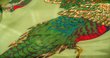 Hareket ve detayı vurgulayan, pürüzsüz yeşil kumaş zemin üzerinde canlı kırmızı, yeşiller ve altınlar ile oluşturulan, uçan iki sülünün dinamik bir tasviri.