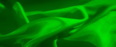 Yeşil kumaştan bir yakın plan, pürüzsüz dokusunu ve doğal kıvrımlarını vurguluyor. Kompozisyon, kumaşın zengin tonunu ve ince ayrıntılarını soyut bir şekilde vurgular.