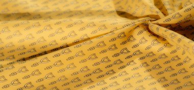 Siyah üzengi desenleri tekrarlayan sarı bir kumaş. Kumaşın yumuşak dokusu ve ince kıvrımları dinamik bir görsel akış yaratır.,