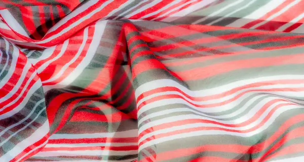 Dinamik kırmızı, beyaz ve yeşil çizgilerden oluşan ipeksi bir kumaşın soyut yakın çekimi. Materyalin yumuşak kıvrımları canlı renkleri ve dokuları vurgulayan yumuşak bir akış yaratır..