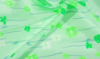 Zarif yeşil kumaşı canlı çiçek desenli, yeşil çiçekli ve açık mavi uzun saplı. Malzemenin akışı ve kıvrımları dinamik, doğadan esinlenilmiş bir tekstil deseni yaratır..