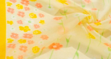 Yumuşak sarı kumaşın bulanık görüntüsü ve çiçekli detaylar. Küçük turuncu ve sarı çiçekler yayılarak hafif, havadar bir doku oluşturur. Kumaşlar nazikçe katlanır akışına