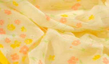 Sarı ve turuncu tonlarda yumuşak çiçek izleriyle süslenmiş narin sarı kumaş bulanıklığı. Kumaşın hafif dokusu, çiçeklerin yumuşak kıvrımlarını ve akan desenlerini yakalar ve rüya gibi bir şekil yaratır.,