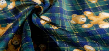 Üzerinde yeşil ve mavi ekoseli, oyuncak ayı resimli ve canlı sarı çiçekli detaylı kumaş görüntüsünü bulanıklaştırın. Dönen kıvrımlar, oyuncu kalıplara dikkat çekiyor.