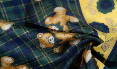 Üzerinde yeşil ve mavi ekoseli, oyuncak ayı resimli ve canlı sarı çiçekli detaylı kumaş görüntüsünü bulanıklaştırın. Dönen kıvrımlar, oyuncu kalıplara dikkat çekiyor.