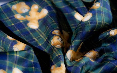 Ekose bir arka planla tamamlanmış, çiçek ve oyuncak ayı motifleri içeren yeşil ve sarı bir kumaş bulanıklığı. Kalıpların birleşimi eşsiz, sanatsal bir görünüm yaratır.