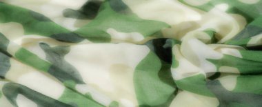 Canlı yeşil ve bej rengi kamuflaj kumaşı, ışık ve gölgenin etkileşimiyle derinlik ve boyut oluşturarak yavaşça akar. Kumaşın yumuşak dokusu huzur ve sükunet hissi uyandırır..