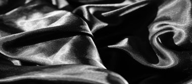 Bulanık siyah kumaş, yumuşak röfleli ve koyu gölgeli lüks bir dokuya sahip akışkan dalgalar halinde dizilmiş. Parlak yüzey, saten maddenin zenginliğini vurgulayan ışığı yansıtıyor.