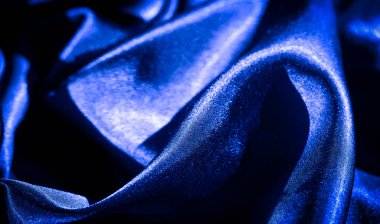 Pürüzsüz, sıvı kıvrımlı parlak mavi saten kumaş. Yumuşak, parlak dokusu ışığı yansıtıyor, lüks, ipeksi materyali vurguluyor ve koyu mavi kumaş boyunca zarif kıvrımlar oluşturuyor..