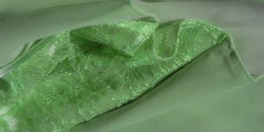 Bulanık yeşil dantel kumaş küçük mor süslemeler ve fiyonklarla süslenmiş, büyüleyici bir kontrast oluşturuyor. Karmaşık ağ tasarımı bu güzel tekstil resmine derinlik katıyor..