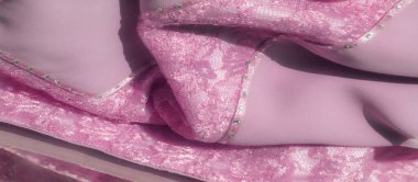 Bulanık pembe dantel kumaş manzarası, ince çiçek detayları ve ızgara örgüsü deseni. Dantel, pırıl pırıl bir kontrast sağlayan, küçük değerli taşlı hassas bir kurdele ile vurgulanır..