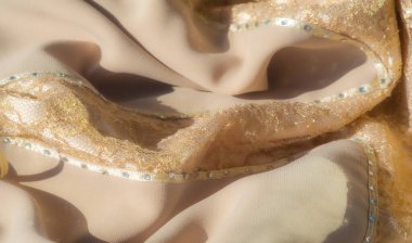 Dantelle sarmalanmış parlak bej kumaş bulanıklığı, hassas kurdeleler ve karmaşık boncuk işçiliğiyle aksanlı. Yumuşak kıvrımlar lüks ve sofistike bir estetik yaratıyor..