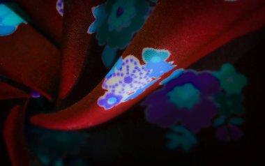 Kırmızı kumaş üzerinde parlak mavi ve mor renkte karmaşık çiçek desenleri. Zıt renklerin kombinasyonu çarpıcı, parlak bir etki yaratır, zengin doku ve akışkan kumaş katları ile..