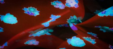 Pürüzsüz kırmızı kumaşta parlayan parlak mavi ve mor çiçek motifleri. Kumaşın kıvrımları derinlik ve doku yaratır, yumuşak maddeye karşı parlak çiçekli tasarımı vurgular..