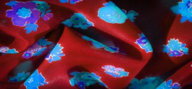 Bulanık kırmızı kumaş açık mavi ve mor çiçek motiflerini gösteriyor. Derin bir arkaplanda parıldıyor. Malzemenin karmaşık kıvrımları yumuşak gölgeler oluşturur ve derinlik ekler.