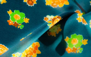 Sarı, turuncu ve yeşil renklerle karışık çiçek desenli bulanık kumaş görüntüsü, güzel bir mavi kumaş arka planının üzerine serilmiş. Yumuşak gölgeler ve kıvrımlar genel tasarıma derinlik ekler.