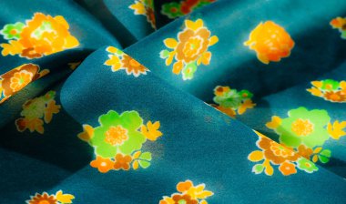 Bulanık, yeşil, turuncu ve sarı çiçekli yumuşak çiçekli kumaş koyu mavi arkaplan üzerinde uyumlu bir denge oluşturuyor. Kumaş kıvrımları ona zarif ve akıcı bir görünüm veriyor..