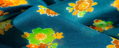 Bulanık görüntü, parlak yeşil, turuncu ve sarı desenleri zengin mavi kumaşa karşı sergiliyor. Doku yumuşak ve narin, üst üste gelen kıvrımlar derinlik ve hareket hissi yaratıyor.