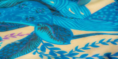 Karmaşık desenli mavi kumaş, bej ve mor renklerde çiçekli ve desenli motifler, yumuşak krem renkli püsküllerle geliştirilmiş, dokunsal ve zengin bir tekstil deneyimi yaratıyor..