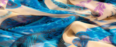 Mavi ve bej tonlarında, karışık mor çiçekli ve sarmaşık desenli bulanık kumaş. Yumuşak püsküllerle süslenmiş kumaşın şeffaf kalitesi ona hafif ve hafif bir his veriyor..