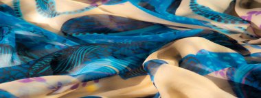 Canlı mavi kumaşın bulanık, narin mor çiçek aksanları ve akıcı bej kıvrımları var. Karmaşık tasarım ve doku görsel olarak dinamik bir tekstil yaratır..