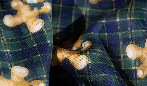 Ekose ve çiçek desenli yeşil bir kumaşın detaylı görüntüsü, büyüleyici oyuncak ayı tasarımları içeriyor. Kontrastlı sarı, tekstile dinamik ve canlı bir dokunuş ekler..