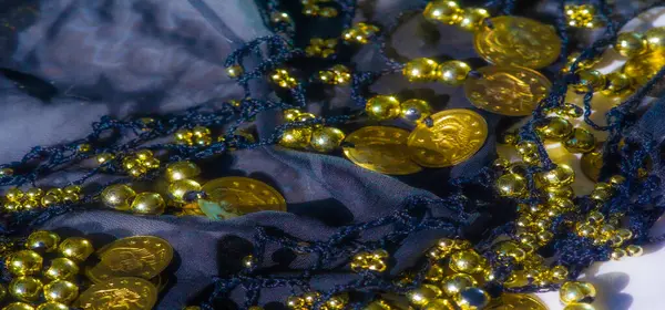 Karmaşık bir desen içinde altın sikkeler ve boncuklar sıralanmış koyu donanma kumaşının yakın plan görüntüsü. Işıldayan altın yumuşak, şeffaf kumaşa karşı duruyor ve zarafetini arttırıyor..
