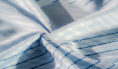 Yumuşak mavi kumaş, hafif dalgalanmalar ve ince çizgili desenler, ışık narin ve pürüzsüz dokuyu güçlendirir, sakin ve zarif bir görsel akış yaratır..