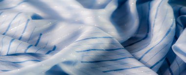 Dikey çizgili narin mavi kumaş bulanıklığı. Işık yüzeyden parıldıyor, ona parıltılı ve akışkan bir görünüm veriyor, kumaşta yumuşak kıvrımlar var..