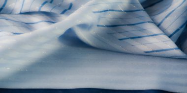 Yüzeyden yansıyan parıldayan ışıkla birlikte mavi kumaştan bulanık, narin kıvrımlı ve ince çizgili desenli yumuşak, akışkan bir görünüm yaratıyor..