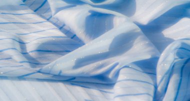 Işıldayan iplikleri ve ince çizgili desenleri olan açık mavi kumaş bulanıklığı. Kumaşın yumuşak, akışkan dokusu ve nazik kıvrımları zarif ve huzurlu bir görsel yaratıyor..