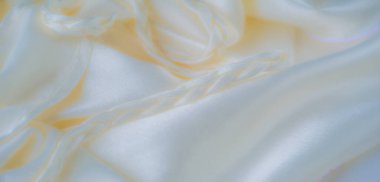 Açık renkli kumaş, örülmüş aksanlar, gölgeler ve dokular etkileşimi yaratarak saten maddenin pürüzsüz ve akıcı doğasını vurguluyor.