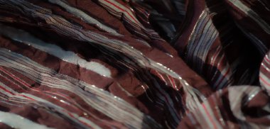 Görüntü. Beyaz ve kırmızı çizgili kahverengi bir kumaş, parıldayan metalik ipliklerle vurgulanıyor, zarif ve yansıtıcı bir yüzey oluşturuyor. Kumaşın yumuşak kıvrımları ve dokulu yüzeyi sunar