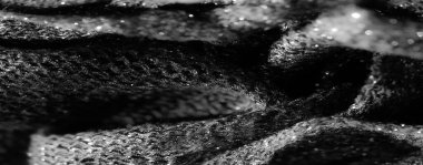 Karanlık, desenli bir kumaşın yakın plan görüntüsü parıldayan bir yüzey ve ince yansıtıcı detaylar sergiliyor, karmaşık dokumasını aydınlatıyor. Malzemenin ince kıvrımları derinlik ekler.