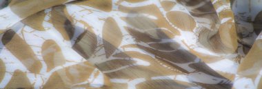 Bulanık. Açık kahverengi bir kumaşın detaylı bir yakın çekimi. Yumuşak, beyaz, soyut yaprak şekilleriyle süslenmiş. Kumaşın yumuşak kıvrımları ve organik ve akışkan bir görünüm oluşturan hassas bir dokusu vardır..
