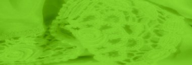 Yumuşak kumaşı kaplayan karmaşık yeşil dantel desenli bulanıklık. Dantel tasarımı, kumaşın pürüzsüz kıvrımlarıyla uyumlu bir şekilde harmanlanarak şık ve zarif bir görünüm sağlar.